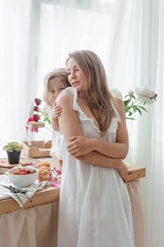 A little blonde girl with her mom on a kitchen countertop decorated with peonies. The concept of the relationship between mother and daughter. Spring atmosphere.