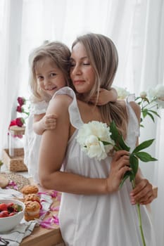 A little blonde girl with her mom on a kitchen countertop decorated with peonies. The concept of the relationship between mother and daughter. Spring atmosphere.