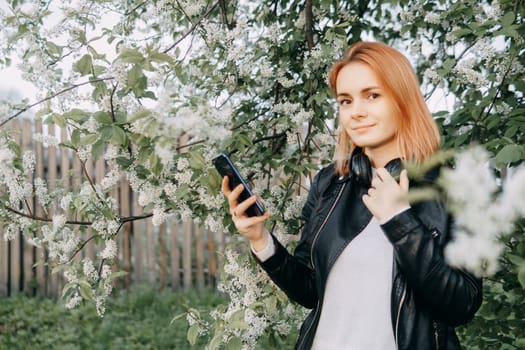 A red-haired teenage girl listens to music in headphones, configured via the phone. The time of spring flowering of cherry orchards.