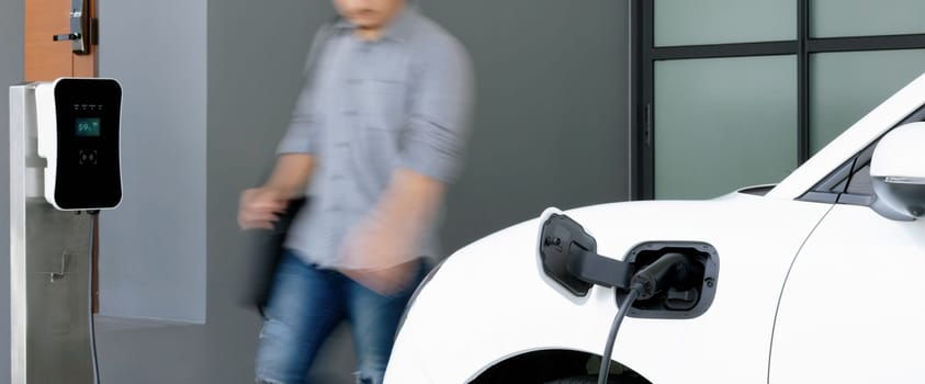 Focus electric car charging at home charging station with blurred progressive man walking in the background. Electric car using renewable clean for eco-friendly concept.