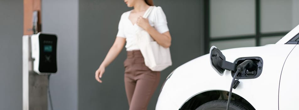 Focus image of electric vehicle recharging battery at home charging station with blurred woman walking in the background. Progressive concept of green energy technology applied in daily lifestyle.