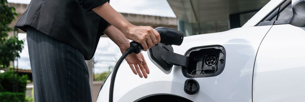 Closeup progressive suit-clad businesswoman with her electric vehicle recharge her car on public charging station in modern city with power cable plug and renewable energy-powered electric vehicle.