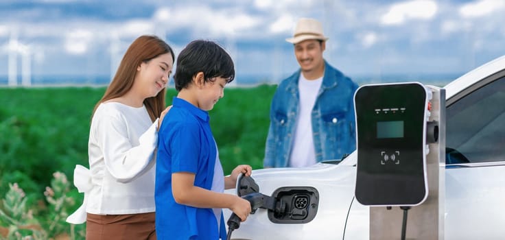 Concept of progressive happy family enjoying their time at wind farm with electric vehicle. Electric vehicle driven by clean renewable energy from wind turbine generator for charging station.