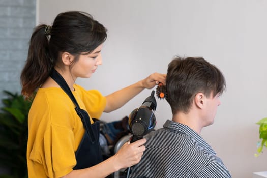Asian female barber making haircut for male customer with male hairstyle in qualified barbershop. Men's hairstyling by a scissor and comb in hair salon concept.