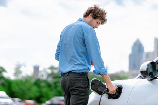 Progressive eco-friendly concept of parking EV car at public electric-powered charging station in city with blur background of businessman leaning on recharging-electric vehicle with coffee.