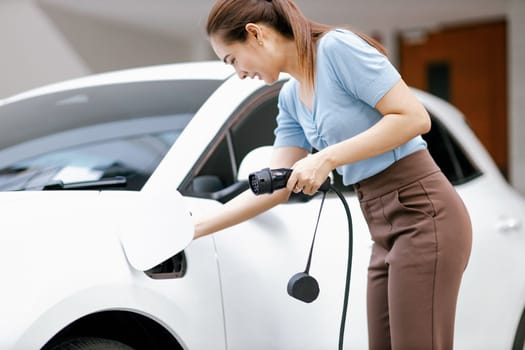 Progressive woman install cable plug to her electric car with home charging station. Concept of the use of electric vehicles in a progressive lifestyle contributes to clean environment.