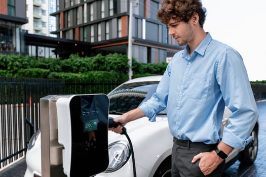 Progressive businessman insert charger plug from charging station to his electric vehicle with apartment condo building in background. Eco friendly rechargeable car powered by sustainable energy.