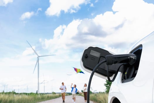 Concept of progressive happy family holding windmill toy and relax at wind farm with electric vehicle. Electric vehicle driven by clean renewable energy from wind turbine generator to charger station.