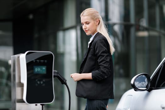 Progressive businesswoman insert charger plug from charging station to her electric vehicle with apartment condo building in background. Eco friendly rechargeable car powered by sustainable energy.