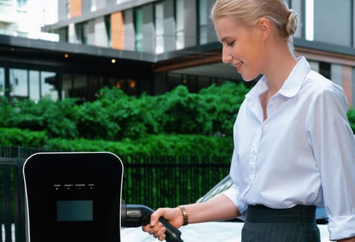 Progressive businesswoman insert charger plug from charging station to her electric vehicle with apartment condo building in background. Eco friendly rechargeable car powered by sustainable energy.