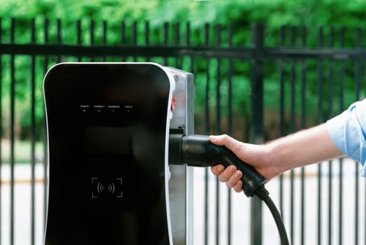 Closeup progressive man holding EV charger plug from public charging station for electric vehicle with background of residential building as concept eco-friendly sustainability energy car concept.