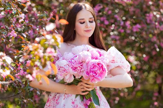 A portrait of a smiling beautiful girl holding a large bouquet of pink peonies in her hands stands with her eyes closed, in a blooming garden on a sunny day. Copy space