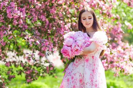 A beautiful girl in a light pink dress, with a large bouquet of pink peonies, stands next to her in a blooming garden on a sunny day. Copy space