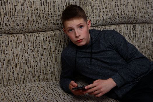 A boy in a sweatshirt lying on the sofa with a smartphone.
