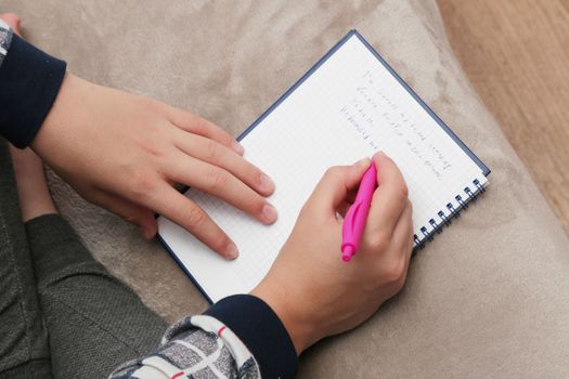 The boy sits on the sofa and makes notes in the notebook.