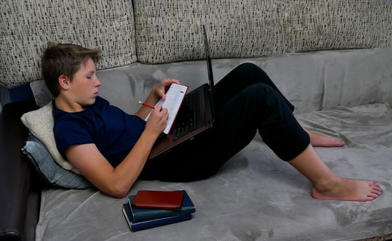 A boy in a blue T-shirt lies on the couch with a laptop and writes homework in a notebook. The teenager is engaged in distance learning with a laptop and notebook. teenager is doing self-study