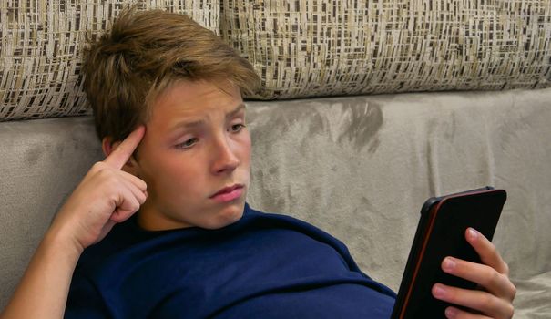 The teenager in a blue T-shirt holds an electronic gadget in his hand and spins his finger at the temple.