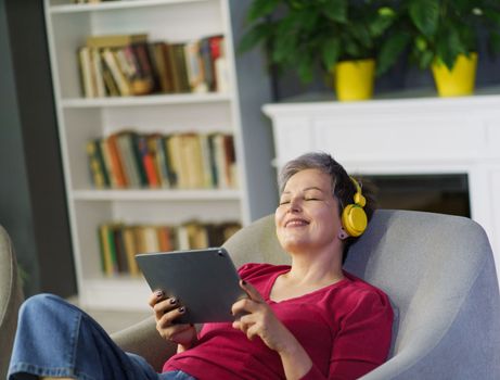 Serene, mature woman with grey hair enjoying her leisure time indoors while listening to music through headphones and holding a tablet in her hands. She appears relaxed and content, displaying the peacefulness. High quality photo