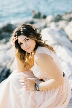 Bride in a beige dress sits on the rocks by the sea with her head propped up by her hand. High quality photo