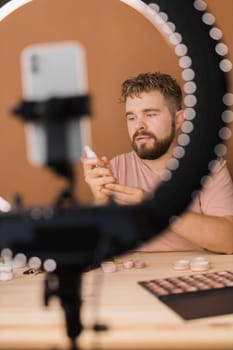Portrait of happy man beauty blogger sitting in room at table and speaking recommending foundation or decorative cosmetic looking at camera. Blogging and vlog