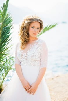 Bride in a white lace dress stands near a palm tree on the seashore. High quality photo