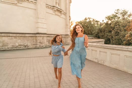 Daughter mother run holding hands. In blue dresses with flowing long hair against the backdrop of a sunset and a white building