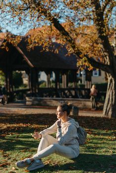 Young fashionable teenage girl with smartphone in park in autumn sitting at smiling. Trendy young woman in fall in park texting. Retouched, vibrant colors. Beautiful blonde teenage girl wearing casual modern autumn outfit sitting in park in autumn. Retouched, vibrant colors, brownish tones.