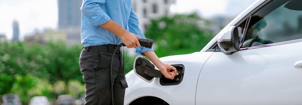 Closeup progressive man holding EV charger plug from public charging station for electric vehicle with background of residential building as concept eco-friendly sustainability energy car concept.