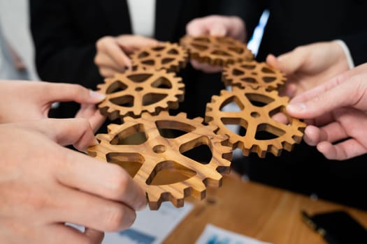 Closeup top view businesspeople hand holding gear and join together over meeting table with financial report papers. Cohesive group of office workers holding cog wheel as synergy harmony concept.