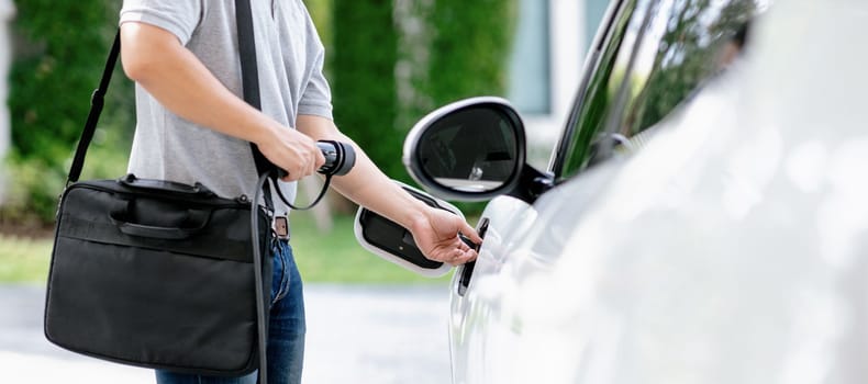 Progressive asian man install cable plug to his electric car with home charging station in the backyard. Concept use of electric vehicles in a progressive lifestyle contributes to clean environment.