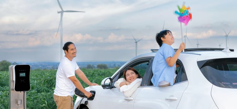 Concept of progressive happy family enjoying their time at wind farm with electric vehicle. Electric vehicle driven by clean renewable energy from wind turbine generator for charging station.