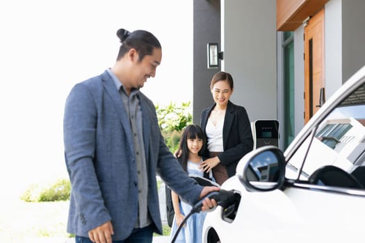 Progressive young parents and daughter with electric vehicle and home charging station. Green and clean energy from electric vehicles for healthy environment. Eco power from renewable source at home.