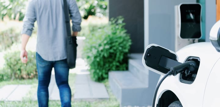 Focus electric car charging at home charging station with blurred progressive man walking in the background. Electric car using renewable clean for eco-friendly concept.