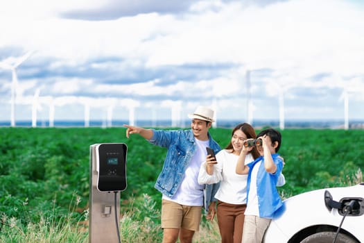 Concept of progressive happy family enjoying their time at wind farm with electric vehicle. Electric vehicle driven by clean renewable energy from wind turbine generator for charging station.