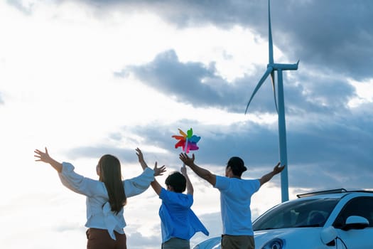 Concept of progressive happy family enjoying their time at wind farm with electric vehicle. Electric vehicle driven by clean renewable energy from wind turbine generator for charging station.