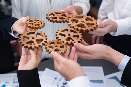 Closeup top view businesspeople hand holding gear and join together over meeting table with financial report papers. Cohesive group of office workers holding cog wheel as synergy harmony concept.