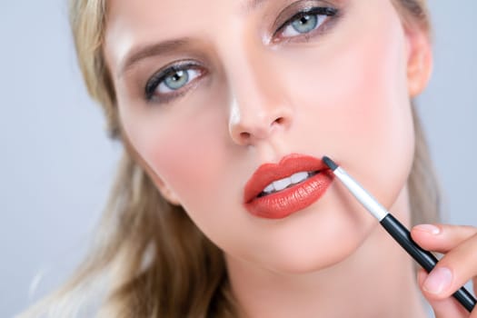 Closeup beautiful young woman with flawless healthy skin and natural makeup putting alluring fashion glossy red lipstick on her lip with lip brush in isolated background.