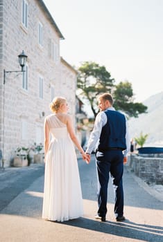 Bride and groom hold hands, standing near the stone house on the street. High quality photo