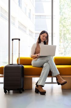 Vertical image of young woman waiting in hotel reception with suitcases. Working with laptop. Lifestyle and travel concept.