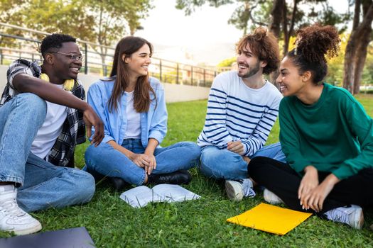 Multiracial college students study together sitting on campus grass. Friends talking in park. Higher education concept.