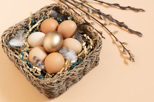 Easter eggs natural and gold in basket with feather. Branch of willow catkins. Light pastel orange background with copy space.