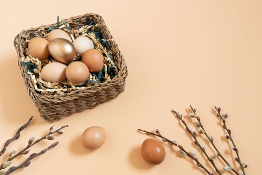 Easter eggs natural and gold in basket with paper filler . Branch of willow catkins. Light pastel orange background with copy space.