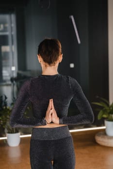 A girl in a black sports uniform does yoga standing on a mat in the gym.