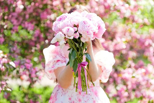 A large bouquet of peonies in the hands of a girl standing next to pink blooming apple trees in the garden, on a sunny day. Front view. Selective focus. Close up. Copy space