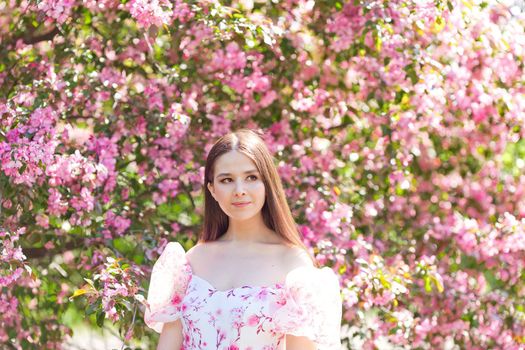 Portrait beautiful brunette girl in light pink dress, is standing under a pink blooming apple trees, in the spring in the garden. Looks to the side. Copy space