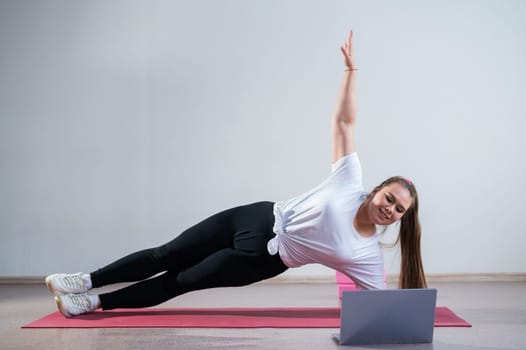 Young fat caucasian woman doing a side plank on a white background. A cute plus size girl in sportswear is doing fitness exercises and watching an online tutorial on a laptop.