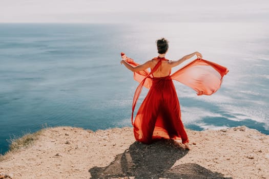 Side view a Young beautiful sensual woman in a red long dress posing on a rock high above the sea during sunrise. Girl on the nature on blue sky background. Fashion photo.