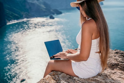 Successful business woman in yellow hat working on laptop by the sea. Pretty lady typing on computer at summer day outdoors. Freelance, travel and holidays concept.