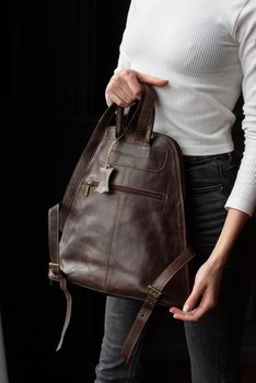 a blond girl in a white shirt poses while standing with a brown leather backpack in her hands