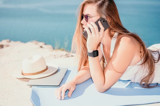 Successful business woman in yellow hat working on laptop by the sea. Pretty lady typing on computer at summer day outdoors. Freelance, travel and holidays concept.